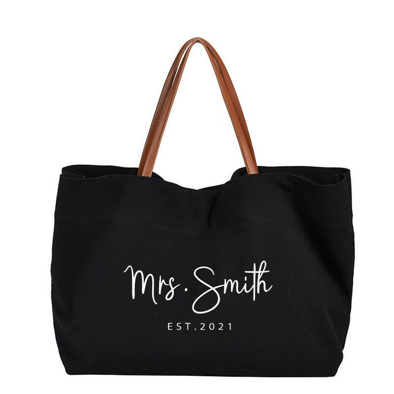 May include: Black canvas tote bag with brown leather handles. The bag has white text that says "Mrs. Smith EST. 2021".