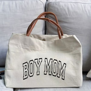 May include: A beige canvas tote bag with brown leather handles. The bag has the text "BOY MOM" printed in black on the front.