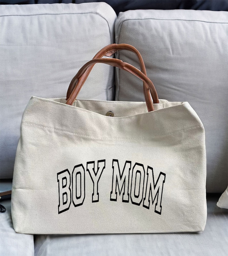 May include: A beige canvas tote bag with brown leather handles. The bag has the text "BOY MOM" printed in black on the front.