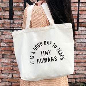 May include: A white canvas tote bag with black text that reads "It is a good day to teach tiny humans".