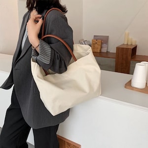 May include: A large, off-white canvas tote bag with brown leather handles. The bag is being held by a person wearing a gray blazer and black pants.