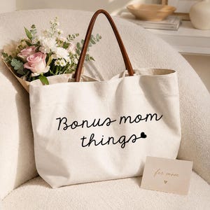 May include: A cream-colored canvas tote bag with brown leather-like handles. The front features the black text 'Bonus mom things' with a small heart icon. A bouquet of pink roses and white flowers is tucked inside, and a small card reads 'for mom'.