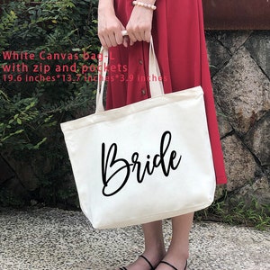 May include: White canvas tote bag with a black "Bride" inscription. The bag has a zipper and pockets and measures 19.6 inches by 13.7 inches by 3.9 inches.