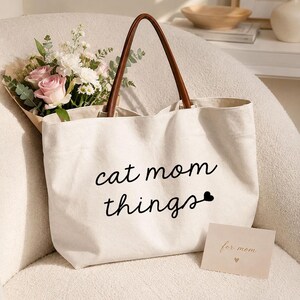 Pode incluir: Uma bolsa tote de lona cor creme com alças castanhas com aspeto de couro. A frente apresenta o texto preto "cat mom things" com um pequeno ícone de coração. Um ramo de rosas cor-de-rosa e flores brancas está dentro, e um cartão diz "for mom".
