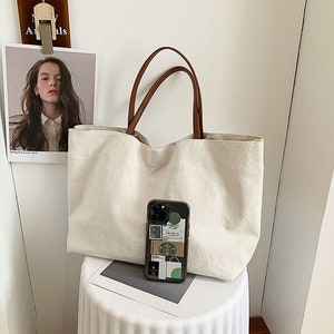 May include: A large, off-white canvas tote bag with brown leather handles. The bag is sitting on a white surface with a mobile phone case with stickers on it.