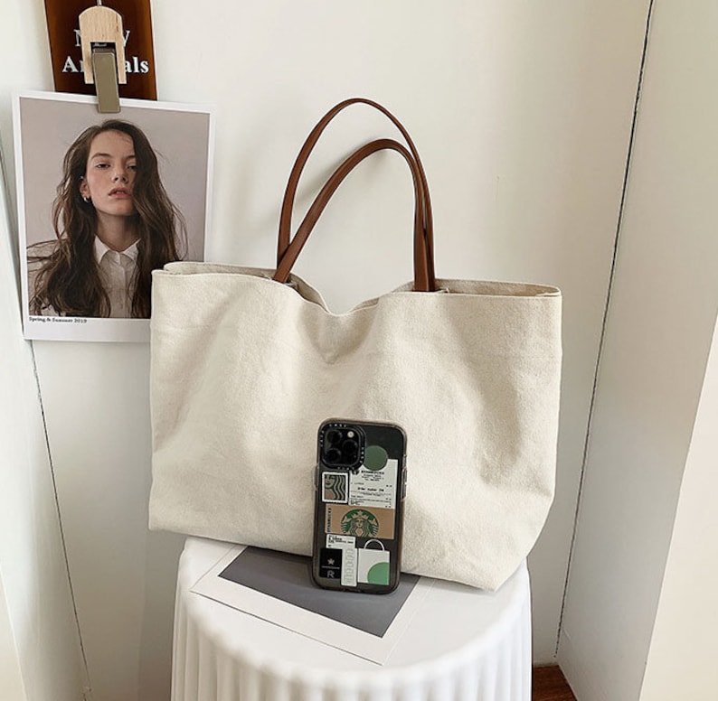 May include: A large, off-white canvas tote bag with brown leather handles. The bag is sitting on a white surface with a mobile phone case with stickers on it.