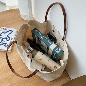 May include: A beige canvas tote bag with brown leather handles. The bag is filled with various items, including a book titled "People's Lawyer: The", a phone, and a water bottle.