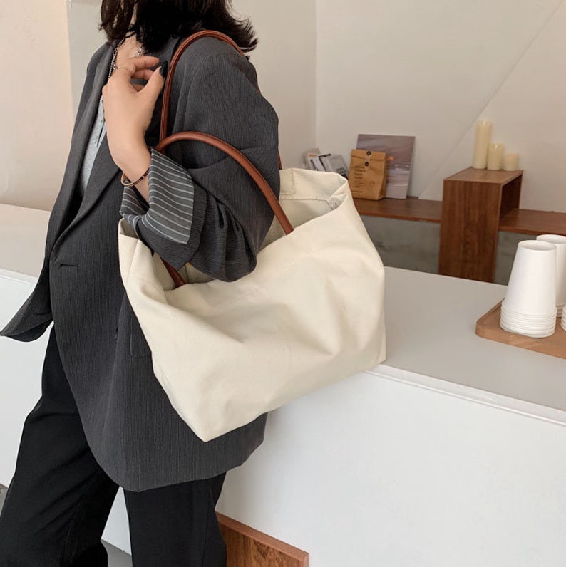 May include: A large, off-white canvas tote bag with brown leather handles. The bag is unzipped and has a relaxed, slouchy look.