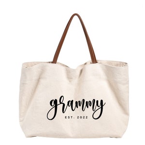 Puede incluir: Una bolsa de lona blanca con asas de cuero marr&oacute;n. La bolsa tiene el texto "grammy est. 2022" impreso en negro.