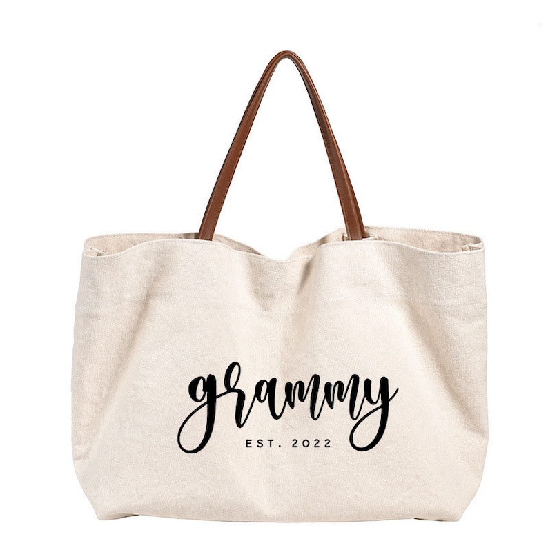 Puede incluir: Una bolsa de lona blanca con asas de cuero marr&oacute;n. La bolsa tiene el texto "grammy est. 2022" impreso en negro.