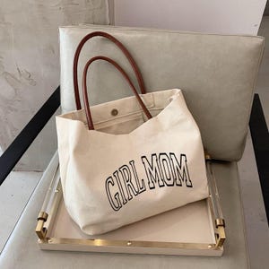 May include: A cream canvas tote bag with brown handles, featuring the black text "GIRL MOM". The bag rests on a tray with gold trim and clear acrylic handles. A light grey chair forms the background.