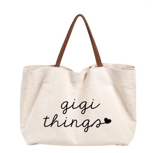 May include: A beige canvas tote bag with brown leather handles. The bag has the text "gigi things" printed on the front in black cursive font with a small black heart.