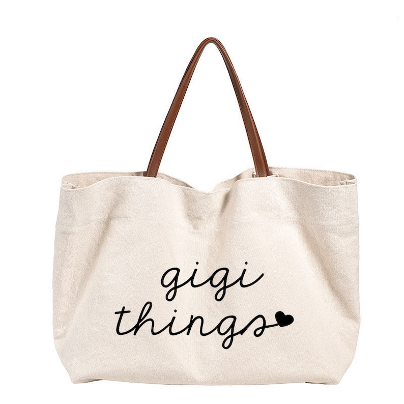May include: A beige canvas tote bag with brown leather handles. The bag has the text "gigi things" printed on the front in black cursive font with a small black heart.