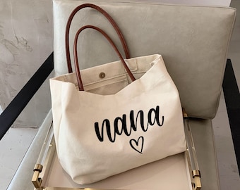 Könnte beinhalten: Eine beige Canvas-Tragetasche mit braunen Ledergriffen. Die Tasche trägt den Schriftzug "nana" in schwarzer Kursivschrift mit einem Herz darunter.