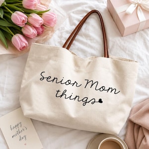 Peut inclure: Un sac fourre-tout en toile de couleur crème avec des anses en similicuir marron. L'avant du sac présente le texte imprimé en noir "senior Mom things" avec une petite icône de cœur à la fin du mot "things" dans une police cursive.