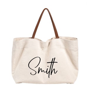 May include: A beige canvas tote bag with brown leather handles. The bag has the word "Smith" written in black cursive on the front.
