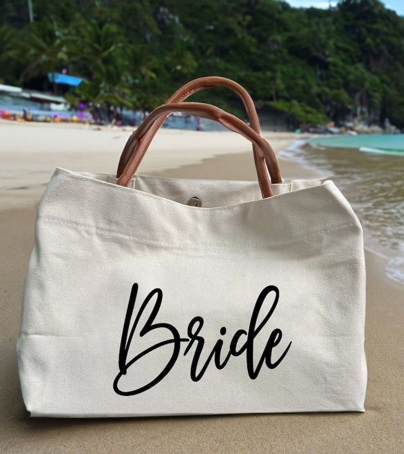 May include: A white canvas tote bag with brown leather handles. The bag has the word "Bride" written in black cursive script.