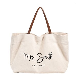 May include: A beige canvas tote bag with brown leather handles. The bag has the text "Mrs. Smith EST. 2021" printed on the front.