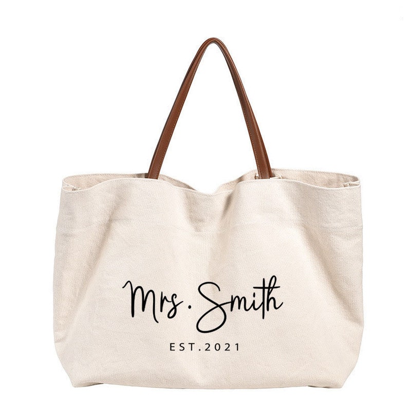 May include: A beige canvas tote bag with brown leather handles. The bag has the text "Mrs. Smith EST. 2021" printed on the front.