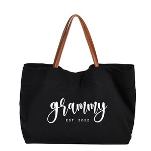 Puede incluir: Bolso tote de lona negra con asas de cuero marr&oacute;n. El bolso tiene el texto "grammy est. 2022" impreso en blanco.