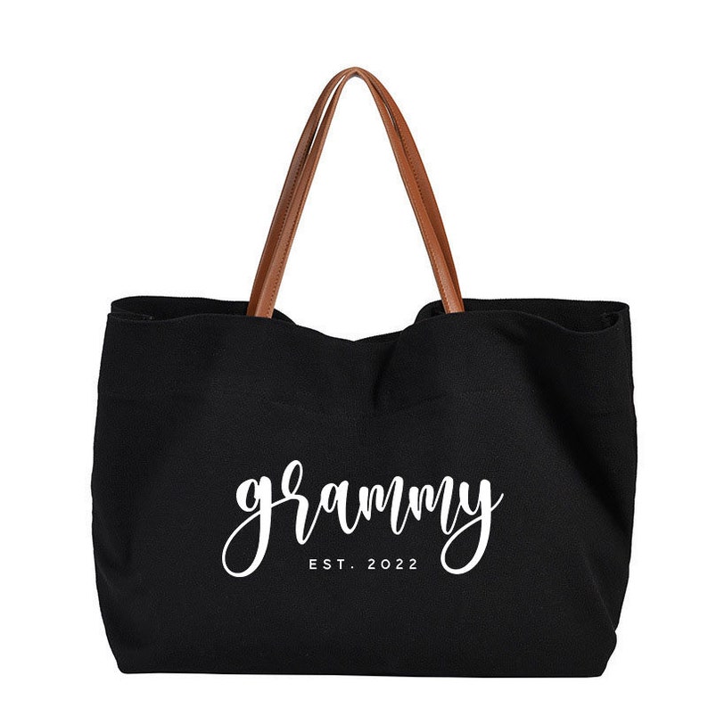 Puede incluir: Bolso tote de lona negra con asas de cuero marr&oacute;n. El bolso tiene el texto "grammy est. 2022" impreso en blanco.
