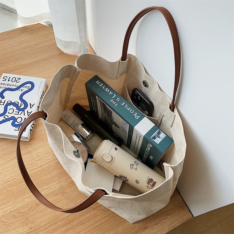 May include: A beige canvas tote bag with brown leather handles. The bag is filled with various items, including a book titled "People's Lawyer: The", a phone, and a water bottle.