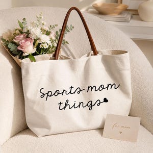 May include: A cream-colored canvas tote bag with brown leather-like handles. The front features the black text "sports mom things" with a small heart icon. A bouquet of pink roses and white flowers is tucked behind the bag, and a small card reads "for mom".