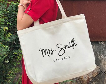 May include: A white canvas tote bag with black text that reads "Mrs. Smith EST. 2021".