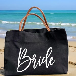 May include: A black canvas tote bag with brown leather handles. The bag has the word "Bride" written in white cursive on the front.