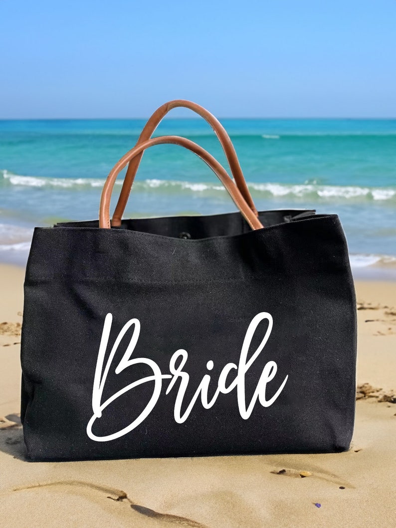 May include: A black canvas tote bag with brown leather handles. The bag has the word "Bride" written in white cursive on the front.