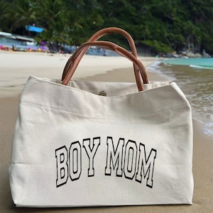 May include: A white canvas tote bag with brown leather handles. The bag has the text "BOY MOM" printed in black on the front.