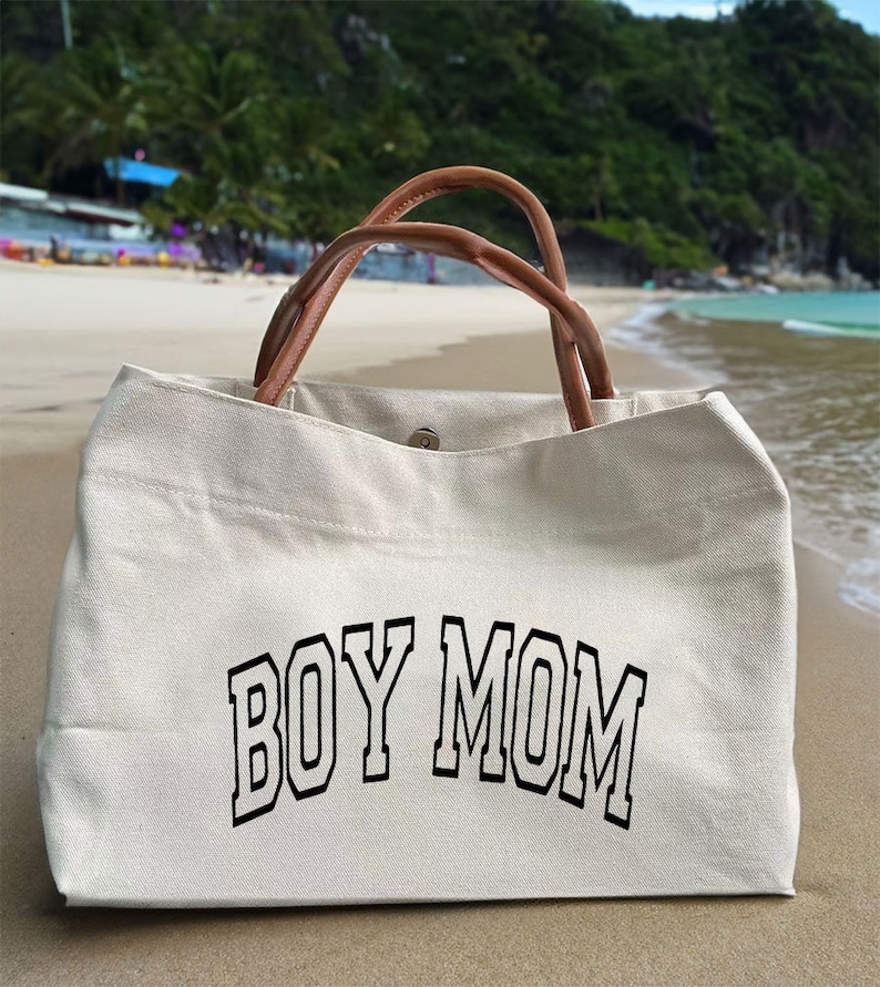 May include: A white canvas tote bag with brown leather handles. The bag has the text "BOY MOM" printed in black on the front.