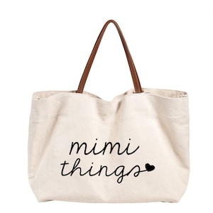 Op de afbeelding: Een beige canvas tote bag met bruine leren handvatten. De tas heeft de tekst "mimi things" in zwarte cursieve letters met een klein zwart hartje.