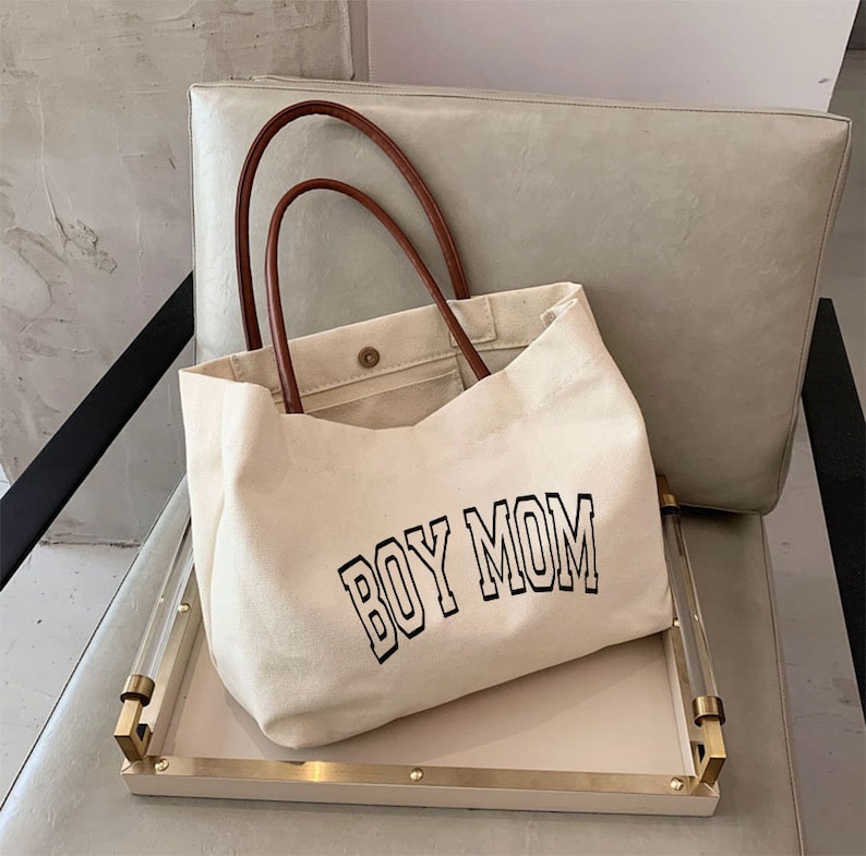 May include: A white canvas tote bag with brown leather handles. The bag has the text "BOY MOM" printed in black on the front.
