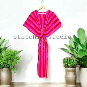 Puede incluir: Un vibrante caftán a rayas rosas y rojas colgado de una percha de madera. El vestido está atado a la cintura y tiene un escote en V. El texto "stitchingstudio" es visible en la pared. El fondo incluye plantas en macetas.