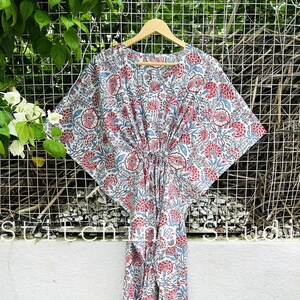 May include: A white, red, and blue floral print kaftan dress hanging on a wooden hanger. The dress features a V-neck and a cinched waist. The fabric is patterned with large floral motifs and smaller leaf designs. The words "stitch studio" are visible at the bottom.