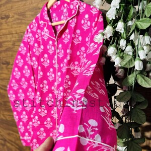 Puede incluir: Conjunto de pijama rosa brillante con un estampado floral blanco y ribete blanco. La parte superior de manga larga y los pantalones se muestran en una percha de madera. El texto "stitchingst" es visible en la tela.