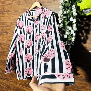 Puede incluir: Una camisa de pijama de manga larga con un estampado de rayas blancas y azul marino. La camisa presenta un diseño repetido de ilustraciones de pavo real rosa. La camisa es abotonada con un bolsillo en el pecho y está colgada en una percha de madera.