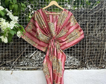 May include: A red and gold striped kaftan dress with a V-neck and a tie at the waist. The dress features a repeating pattern of tigers. The garment is hanging on a wooden hanger against a white wall and a wire mesh background.