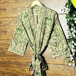 May include: A light green floral print robe with a white trim, hanging on a wooden hanger. The robe features a V-neck and a tie closure at the waist. The floral pattern includes pink flowers and green leaves.