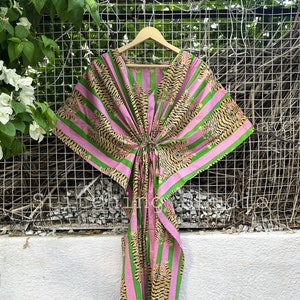 May include: A pink and green striped kaftan dress with a tiger print. The dress is hanging on a wooden hanger against a white wall and a metal grid. The kaftan has a V-neck and a tie at the waist.
