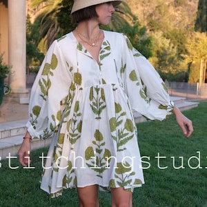 Op de afbeelding: Witte mini-jurk met groene bloemenprint en lange mouwen. De jurk heeft een V-hals en een losse pasvorm. Een strohoed en roze sandalen maken de look compleet. De tekst "stitchingstudio" is zichtbaar.