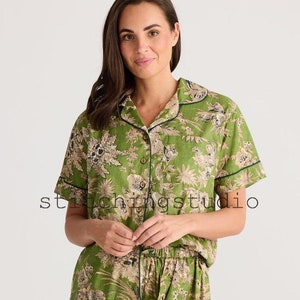 May include: Green floral print pajama set. The set includes a short-sleeved button-down shirt and matching shorts. The edges are trimmed with navy blue piping. The fabric features a botanical design in shades of green and beige.