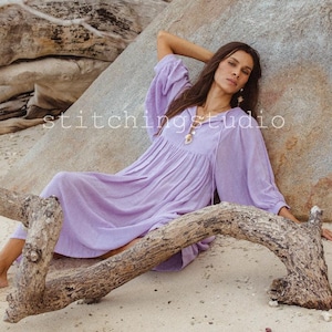 Może przedstawiać: Kobieta w liliowej sukience z długimi, pękatymi rękawami, odpoczywająca na skale i drewnie wyrzuconym na piaszczystej plaży. Sukienka ma luźny, zwiewny krój. Na skale widoczny jest napis "stitchingstudio".