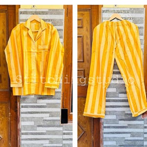 May include: Yellow and white striped pajama set. The set includes a long-sleeved button-down shirt with a chest pocket and matching pants. The shirt and pants are hanging on a wooden hanger. The text "Stitchingstudio" is visible on the shirt.