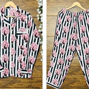 Puede incluir: Un conjunto de pijama a juego con una camisa con botones y un pantalón. El conjunto es de rayas blancas y negras con un diseño de pavo real rosa repetido. La camisa tiene un bolsillo en el pecho y se muestra en perchas de madera.