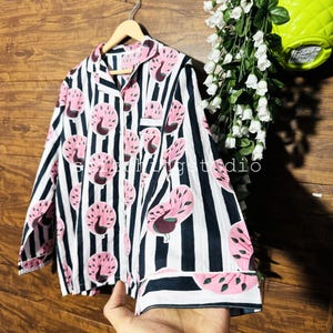 Puede incluir: Una camisa de pijama a rayas blancas y azul marino, adornada con motivos de pavo real rosa. La camisa tiene un bolsillo en el pecho y ribetes azul marino en los puños y el cuello. Cuelga de una percha de madera.