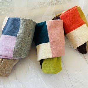 May include: Three pairs of colorful socks with a patchwork design. Each sock features a different color combination, including blue, gray, purple, pink, green, and orange. The socks are folded over, revealing the contrasting colors and patterns.
