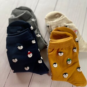May include: Three pairs of socks with penguin designs. One pair is navy blue with white penguins, one pair is mustard yellow with white penguins, and one pair is gray with white penguins.