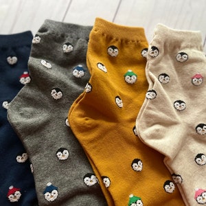 May include: Four pairs of socks with a penguin pattern. The socks are in navy blue, gray, mustard yellow, and beige.